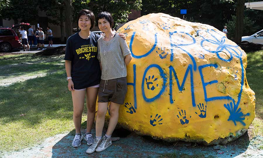 Two women, a student and her parent, pose by a large rock inscribed with the word UR-home at the University of Rochester.